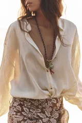 Blusa color crema de tejido arrugado con hilo metalizado, cuello redondo con escote en pico, manga larga abullonada y nudo delantero. Cierre frontal con botones con flores bordadas.
