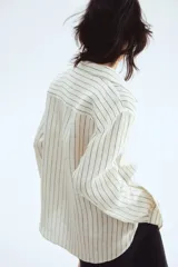 Camisa de lino color blanco con rayas verticales negras, cuello clásico, cierre frontal con botones, manga larga con puños abotonados, bolsillo de parche en el pecho y bajo redondeado.