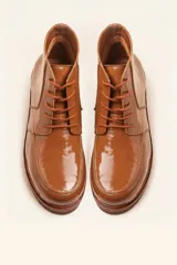 Bota de cuero vacuno con acabado charol, color marrón oscuro. Presenta un diseño de caña corta con cordones frontales y costuras decorativas en la puntera. La suela es de goma tipo crepe.