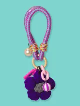 Llavero con cordón elástico celeste y rosa, adornado con una cuenta perlada, una flor tejida violeta con centro gris, una cuenta rayada blanca y violeta, un dije rosa con forma de pastilla y eslabones de cadena rosa.