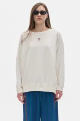 Buzo color crema de corte oversized, con mangas largas, hombros caídos y cuello redondo. Presenta un bordado pequeño en el frente y un bordado grande en la espalda, ambos a tono.