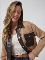 Chaqueta de corte boxy confeccionada en tejido de punto color crema, con detalles en cuero sintético negro en cuello, solapas, cartera central y bolsillos frontales. Presenta cierre frontal con botones metálicos plateados y costuras decorativas visibles.