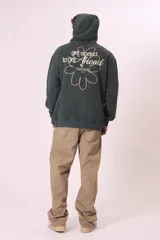 Sudadera con capucha de color verde oscuro (o verde petróleo) con efecto lavado, corte holgado y bolsillo canguro. Presenta un pequeño bordado blanco en el pecho con un diseño floral y texto.