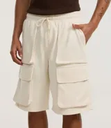 Bermuda baggy color crema, confeccionada en algodón y poliéster. Cuenta con cintura elástica ajustable con cordón y bolsillos cargo laterales.