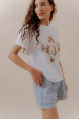 Remera blanca de algodón con calce relajado y estampa frontal de una flor grande con pétalos en tonos neutros y detalles en dorado.