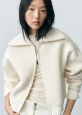 Abrigo corto color crudo de tejido bouclé con mezcla de lana. Tiene cuello de solapa ancha, cierre delantero con cremallera, manga larga con puños elásticos y terminaciones de canalé en contraste. Diseño oversize.