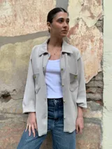 Camisa color beige de tejido suave, con cuello clásico, cierre frontal con botones dorados, mangas largas con puños abotonados y bolsillos de parche en el pecho con botón dorado. Corte holgado.