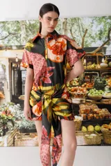 Vestido camisero corto con estampado abstracto en tonos naranja, rosa, amarillo y negro. Tiene cuello camisero, mangas cortas y cierre frontal con botones. Incluye lazo en la cintura para ajustar.