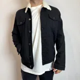 Chaqueta negra de denim con cuello de corderito blanco, cierre frontal con botones y bolsillos en el pecho.