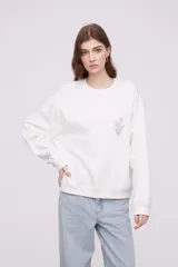 Buzo blanco de corte holgado con cuello redondo y mangas largas. Presenta un pequeño estampado de una flor en el centro del pecho.