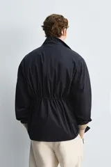 Parka azul marino de tejido técnico, corte relaxed fit, cuello alto ajustable con cordón, manga larga con puños abotonados, cierre frontal con cremallera oculta y solapa con botones a presión, cintura ajustable con cordón interior y bolsillos frontales multifunción de plastrón.