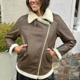 Campera gamulán color marrón con interior de piel sintética color crema, cierre frontal con cremallera descentrada, cuello con solapas amplias y bolsillos laterales.