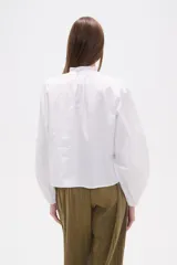 Camisa blanca de algodón con mangas largas abullonadas y cuello mao con volado. Presenta un canesú con detalles de encaje calado en el frente.