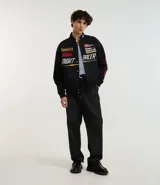 Campera negra con cuello alto, cierre de botones y estampado estilo racing con logos y textos en amarillo, rojo y blanco. Tiene bolsillos laterales y mangas largas con detalles estampados. Marca Blue Steel.