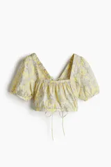 Blusa cropped de algodón color amarillo con estampado paisley en tonos grises. Tiene escote cuadrado elástico, mangas cortas con puños ajustados, espalda descubierta y tiras para atar.