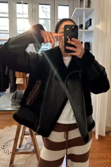 Campera gamulán negra con interior de piel sintética, cierre cruzado con cremallera, cuello con solapas amplias y bolsillos laterales con cremallera. Presenta detalles de cuero en los hombros y un bolsillo con cierre en el pecho.
