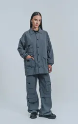 Campera gris de tejido liviano, con cuello camisero, cierre frontal de broches y puños elásticos. Presenta múltiples bolsillos cargo en el frente con ribetes reflectantes blancos.