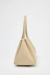 Bolso tipo tote color beige, con doble asa corta y cierre superior con botón imantado. Incluye un compartimento interno con cierre de cremallera.