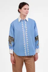 Camisa celeste a rayas verticales blancas, de corte holgado, con cuello clásico blanco, cierre frontal con botones y estampado de leopardo en los puños.