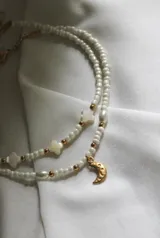 Collar doble de cuentas blancas con detalles dorados, dijes de perlas y dijes de estrellas. El collar inferior tiene un dije de luna dorada.