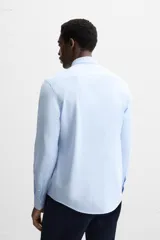 Camisa celeste a rayas verticales blancas, de corte recto, con cuello italiano abierto, manga larga con puños abotonados y cierre frontal con botones.