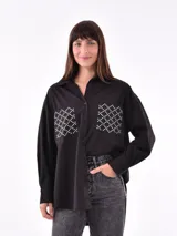 Camisa negra de corte recto, con cuello clásico, cierre frontal con botones y mangas largas con puños abotonados. Presenta dos bolsillos de parche en el pecho con apliques de tachas blancas formando un patrón geométrico.