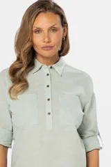 Camisa color beige de lino, de manga larga con botones metálicos personalizados y presilla en la manga. Tiene cuello clásico, cierre frontal parcial con botones y bolsillo de parche en el pecho.