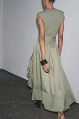 Vestido midi color verde oliva, de cuello redondo y sin mangas. Presenta un cinturón con hebilla metálica y detalle de volantes en la falda.