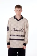 Camiseta oversize de poliéster color crema con diseño a cuadros, cuello en V con ribete negro, mangas largas con puños negros, logo "Blkmba" en el pecho y escudo "Mamba Soccer" en la manga.