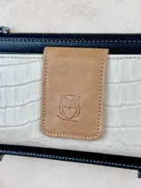 Billetera de cuero con cierre superior y correa de muñeca. Presenta un bolsillo frontal con textura símil cocodrilo y un aplique con el logo de la marca.