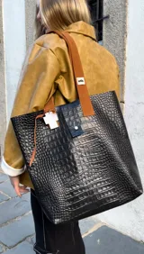 Bolso tote negro con textura símil piel de cocodrilo, doble asa de cuero color marrón y cierre con botón a presión.