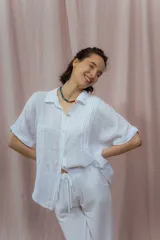 Camisa color crudo confeccionada en lino, de corte holgado, con cuello clásico, mangas cortas con dobladillo y cierre frontal con botones. Presenta detalle de pliegues verticales en la parte frontal.