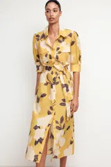 Vestido midi camisero color amarillo con estampado de flores blancas y hojas marrones. Tiene cuello solapa, mangas 3/4 con puños, cinturón de lazo ajustable y cierre frontal con botones.