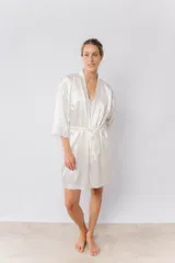 Bata kimono de satén elastizado color blanco, con mangas al codo terminadas en puntilla y largo por encima de la rodilla. Incluye lazo de satén para ajustar en la cintura.