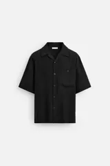 Camisa negra relaxed fit confeccionada en tejido fluido con mezcla de viscosa, cuello bowling, manga corta, bajo con aberturas laterales y cierre frontal de botonadura.