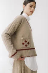 Sweater beige con cuello en V y mangas largas con diseño trenzado. Tiene dos bolsillos frontales con bordados de tréboles rojos y ribetes rojos en cuello, puños y ruedo.