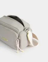 Bandolera gris claro de tela deportiva con bolsillo frontal con cierre y tag de goma. Tiene correa regulable desmontable, acceso mediante cierre superior y bolsillos laterales con cierre y pullers de cordón y goma.