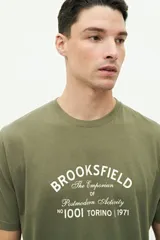 Remera verde militar de manga corta con estampa frontal con el texto "Brookfield The Emporium of Postmodern Activity No 1001 Torino | 1971".
