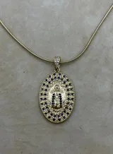 Collar con cadena tipo serpiente dorada y un dije ovalado grande con la imagen de la Virgen de Guadalupe en relieve. El dije está engastado con pequeños cristales azules alrededor del borde y en el centro de la figura. La argolla de sujeción tiene incrustaciones de cristales blancos.