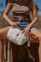 Bandolera color blanco con estampado de manchas grises, correa de cuero marrón y flecos decorativos.