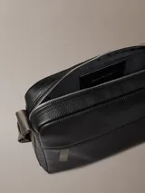 Bolso bandolera rectangular de Calvin Klein, color negro, con textura de cuero granulado. Posee un bolsillo frontal horizontal con cierre y el logo CK grabado en la parte inferior derecha. La correa es ancha y de tela en tono gris/verde oliva, ajustable con hebilla metálica.