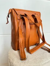 Mochila color marrón de cuero sintético, con asa de mano y correas ajustables. Tiene un bolsillo frontal con cierre a presión y un bolsillo lateral con cierre.