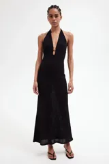 Vestido largo de punto pointelle negro con estilo crochet. Modelo entallado con escote halter en V y adorno de metal en forma de sol en el centro del busto. Falda sirena con bajo recto.