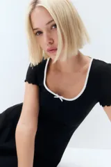 Vestido mini negro de manga corta en punto de algodón acanalado con pollera evasé. Modelo con cuerpo entallado y escote redondo amplio con ribete fino blanco y moña decorativa de satén. Ribete sobrehilado en mangas y bajo.