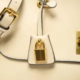 Cartera de mano y hombro color beige, con solapa frontal que se cierra con un herraje metálico dorado. Presenta dos asas superiores y una correa larga desmontable y regulable. Incluye un dije colgante con candado dorado.