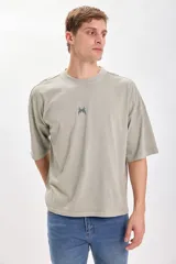 Remera verde de corte amplio y recto, con estampado en el centro de la espalda que incluye el texto "Live Concert" y una lista de géneros musicales.