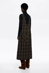 Vestido tipo pichi de largo midi y corte recto, confeccionado en mezcla de lana con diseño a cuadros en tonos verdes y marrones. Presenta escote recto con tirantes anchos ajustables mediante hebillas metálicas, bolsillos de plastrón frontales y traseros, y botones laterales.