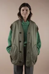 Chaleco largo acolchado color verde oliva, reversible con un lado de piel sintética y otro de pelo sintético. Tiene cuello en V, cierre con botones a presión y bolsillos laterales. Silueta amplia y relajada.