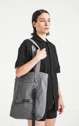 Bolso tipo tote bag color gris, confeccionado en microfibra técnica de secado rápido. Cuenta con un compartimento principal con elástico ajustable, un bolsillo interno con cierre y un bolsillo externo con velcro.