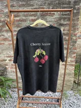 Remera negra con efecto gastado, corte holgado y estampa trasera con la frase 'Cherry Season' y un diseño de cerezas pixeladas.